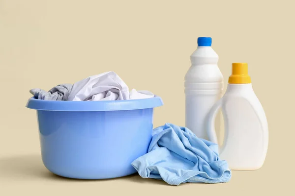 Plastic basin with dirty clothes and bottles of laundry detergent on beige background