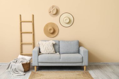 Yastıklı rahat gri kanepe, sepetli ekose, merdiven ve bej duvarın yanında asılı şapkalar.