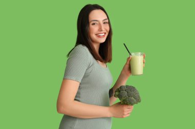 Yeşil arka planda bir bardak smoothie ve brokoli ile genç hamile kadın.