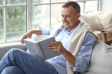 Evde boş gününde kitap okuyan olgun bir adam.