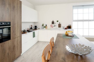 Beyaz tezgahları, dolapları, entegre fırını ve ahşap ada masası olan aydınlık mutfağın içi.