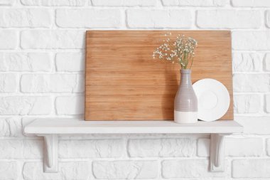 Mutfak rafında kesim tahtası, tabak ve vazo beyaz tuğla duvarda asılı.