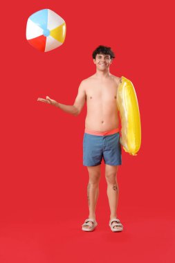 Kırmızı arka planda plaj topu ve yüzme yüzüğü olan genç adam.