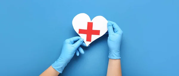 Doctor's hands with paper heart and cross on blue background, top view. Cardiology concept
