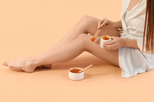 Young woman applying sugaring paste onto her legs against beige background
