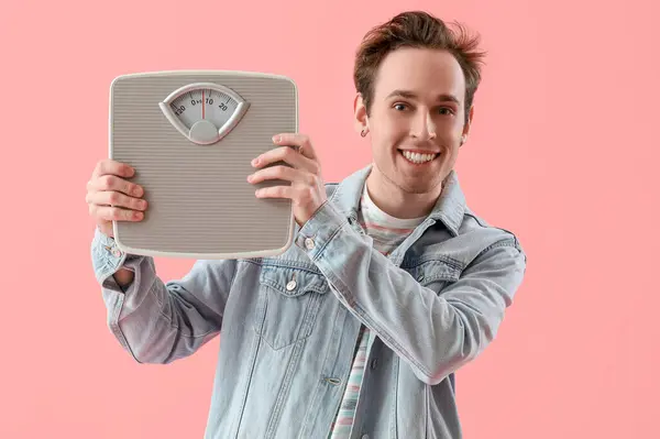 Happy young man with weight scales on pink background