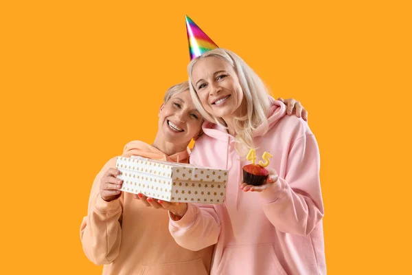 Mature women with birthday muffin and gift on yellow background