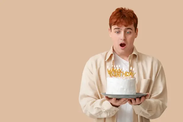 Surprised young man with birthday cake on beige background