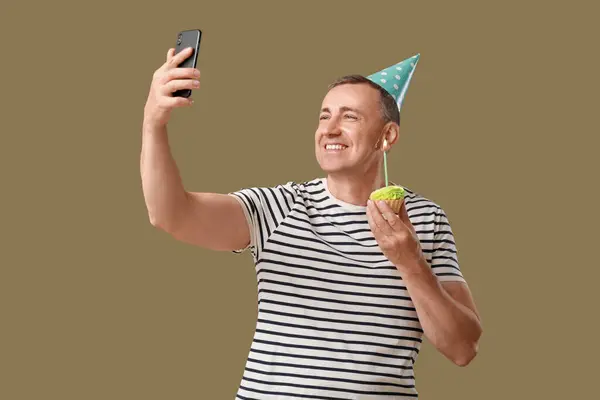Mature man with birthday tartlet taking selfie on green background
