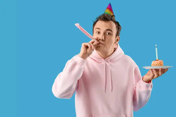 Young man with birthday cake and party whistle on blue background