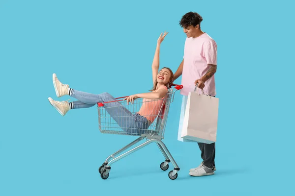 Young man with bags and his girlfriend in shopping cart on blue background