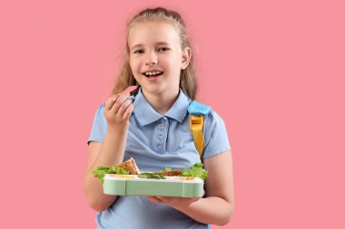 Sırt çantalı ve beslenme çantalı mutlu küçük kız pembe arka planda sebze yiyor.