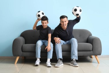 Kızgın küçük çocuk ve babası, mavi duvarın yanındaki kanepede maç izliyorlar.