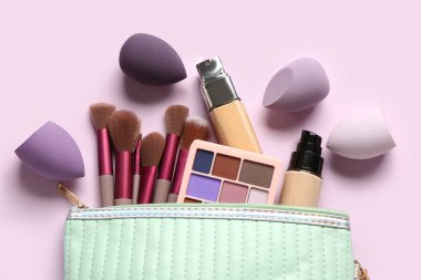 Pembe arka planda farklı makyaj fırçaları, süngerler ve kozmetik ürünleri olan çanta, yakın plan.