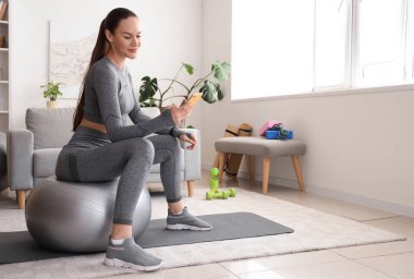 Sportif genç bayan evde fitball oynarken cep telefonu kullanıyor.