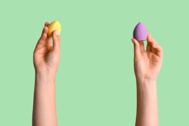 Yeşil arka planda farklı makyaj süngerleri olan kadın elleri