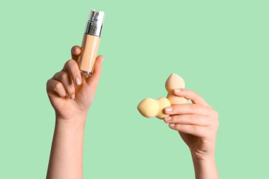 Makyaj süngeri ve yeşil arka planda bir şişe fondöten olan kadın elleri.