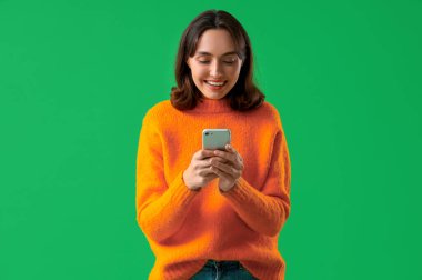 Yeşil arka planda cep telefonu kullanan turuncu kazaklı genç bir kadın.