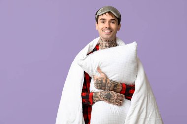 Mor arka planda yastıklı ve battaniyeli pijamalı dövmeli genç adam.