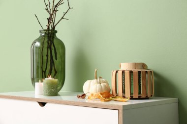 Yeşil duvarın yanında beyaz çekmeceler, balkabakları, mumlar ve ağaç dalları