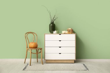 Yeşil duvarın yanında beyaz çekmeceler, balkabakları, mumlar ve ağaç dalları