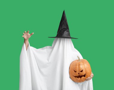 Cadılar Bayramı kostümlü kadın yeşil arka planda balkabağı tutan hayalet ve cadı şapkası.