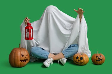 Cadılar Bayramı kostümlü kadın, balkabağı ve fenerle yeşil arka planda oturuyor.