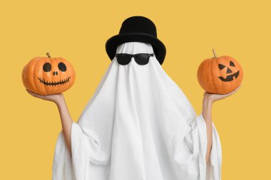 Cadılar Bayramı kostümlü kadın hayalet, silindir şapka ve güneş gözlüğü renkli arka planda balkabağı tutuyor.