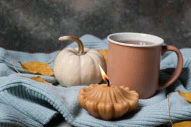 Balkabağı şeklinde yanan mum, bir kupa çay ve gri arka planda sonbahar yapraklarıyla güzel bir kompozisyon.
