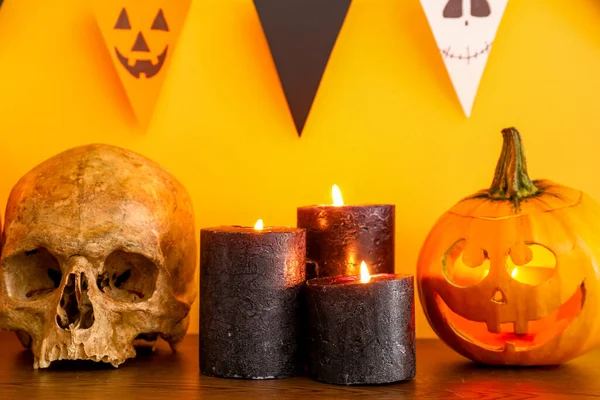 Table with burning candles, pumpkin, skull for Halloween celebration and flags hanging on orange wall in room, closeup