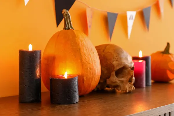 Table with burning candles, pumpkins, skull for Halloween celebration and flags hanging on orange wall in room, closeup