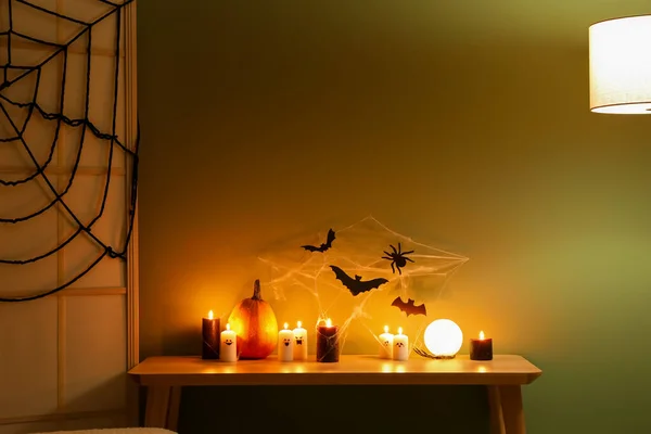 Table with burning candles, lamp, folding screen and Halloween decor near green wall in dark room