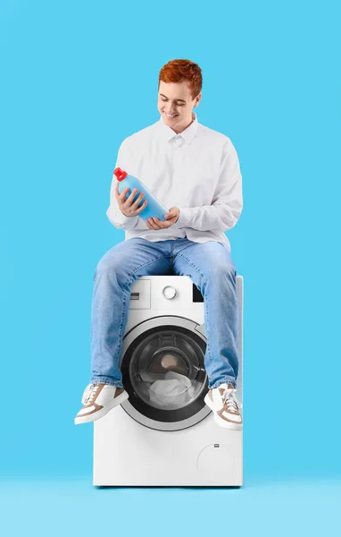 stock image Young man with detergent sitting on washing machine against blue background