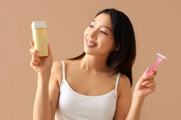 Young Asian woman with liposoluble wax cartridge and razor on beige background