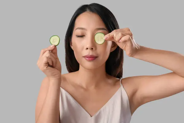 Beautiful Asian woman with cucumber slices on grey background, closeup