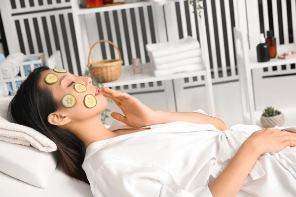Young Asian woman with cucumber slices relaxing in spa salon