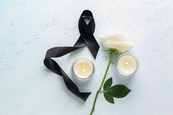 Beautiful rose flower, burning candles and black funeral ribbon on blue background