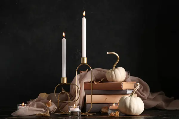Burning candles, scarf, pumpkins, books and autumn leaves on table near black wall