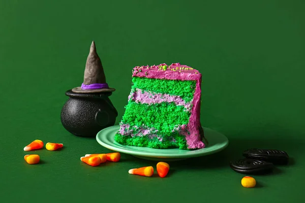 Piece of tasty Halloween cake with candy corns and witch hat on green background