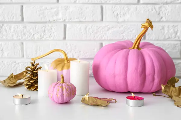 Painted pumpkins with autumn leaves and burning candles on table near white brick wall