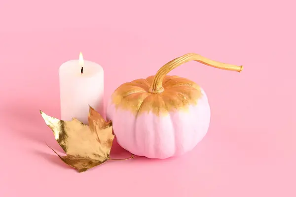 Painted pumpkin with golden leaf and burning candle on pink background