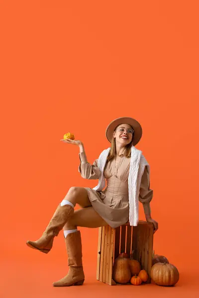 Beautiful young woman with pumpkins and wooden box on orange background