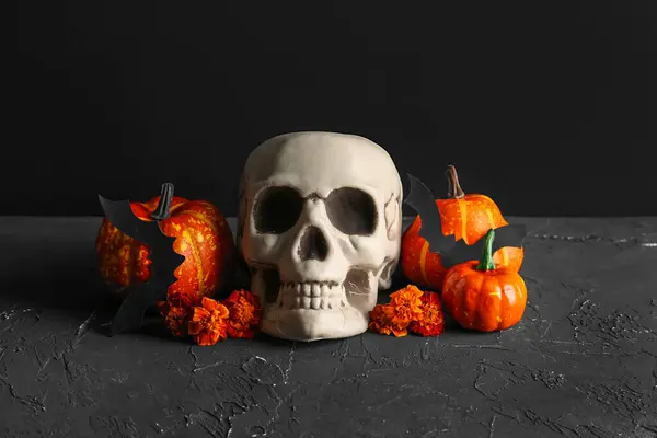 Human skull with marigold flowers, pumpkins and bats on grunge table near black wall