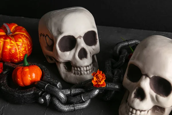 Human skulls with marigold flowers, pumpkins and chain on grunge table near black wall