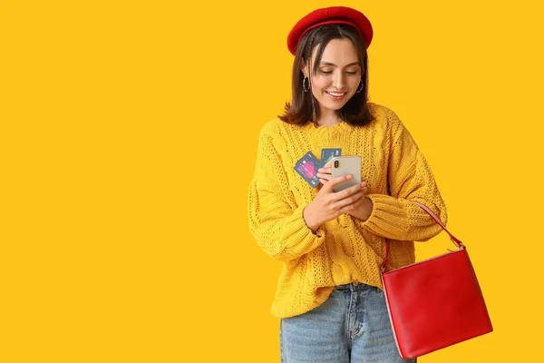 Young woman with credit cards, mobile phone and stylish bag on yellow background