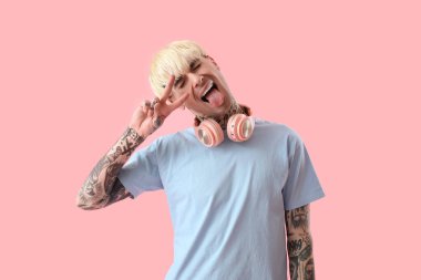 Dövmeli genç adam pembe arka planda zafer işareti gösteriyor.