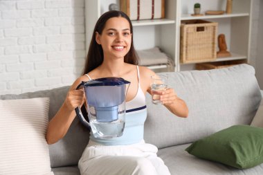 Su filtresi ve cam sürahi ile güzel bir kadın oturma odasında kanepede oturuyor.