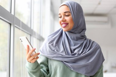 Evin penceresinin yanında cep telefonu kullanan şık bir Müslüman kadın.