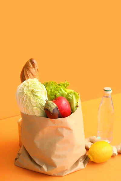 Paper bag with different fresh vegetables, fruits and bottle of water on orange background