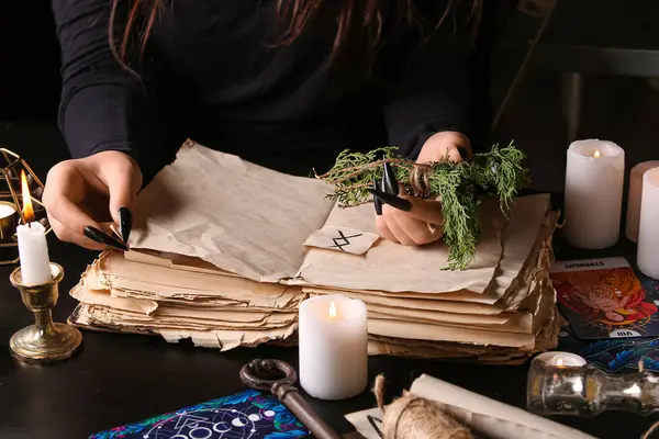 Witch with spell book and herbs at dark table, closeup